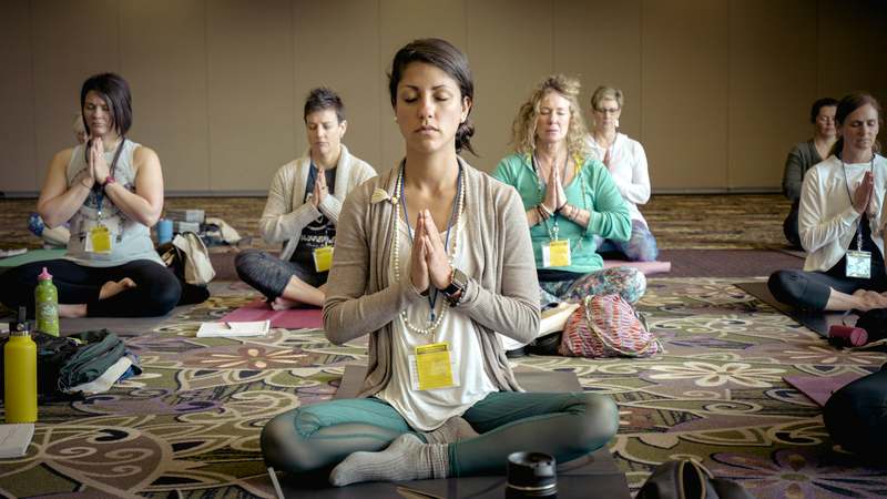 Woman sitting comfortably on a yoga mat beside a window, eyes closed, wearing headphones — peaceful expression, natural lighting